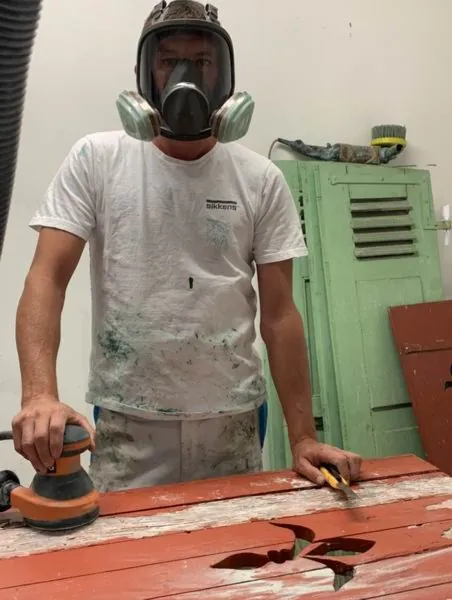 Ponçage d’un volet bois à la machine, artisan protégé par un masque